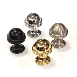 May include: Four decorative cabinet knobs in different finishes: black, chrome, brushed nickel, and gold. Each knob features a spherical top with horizontal grooves and a pedestal base. The knobs are arranged on a white surface.