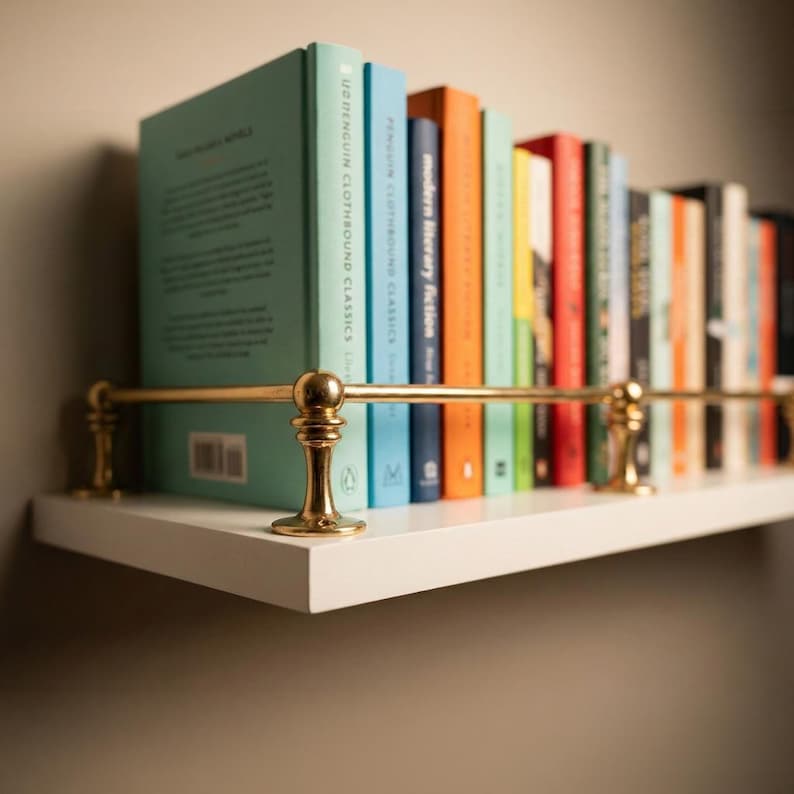 May include: A white shelf with a brass rail holds a collection of colorful books. The books have various colored spines, including teal, blue, orange, and red. The shelf is mounted on a neutral-colored wall, creating a simple, elegant display.