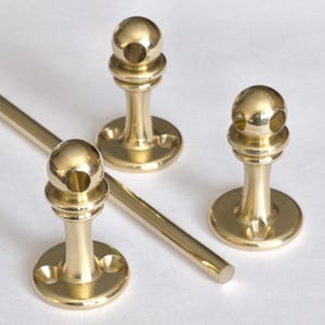 May include: Three brass-colored curtain rod holders and a section of a rod. The holders have a round base, a cylindrical stem, and a spherical top with a hole. The rod is cylindrical and has a polished finish. The items are on a white surface.