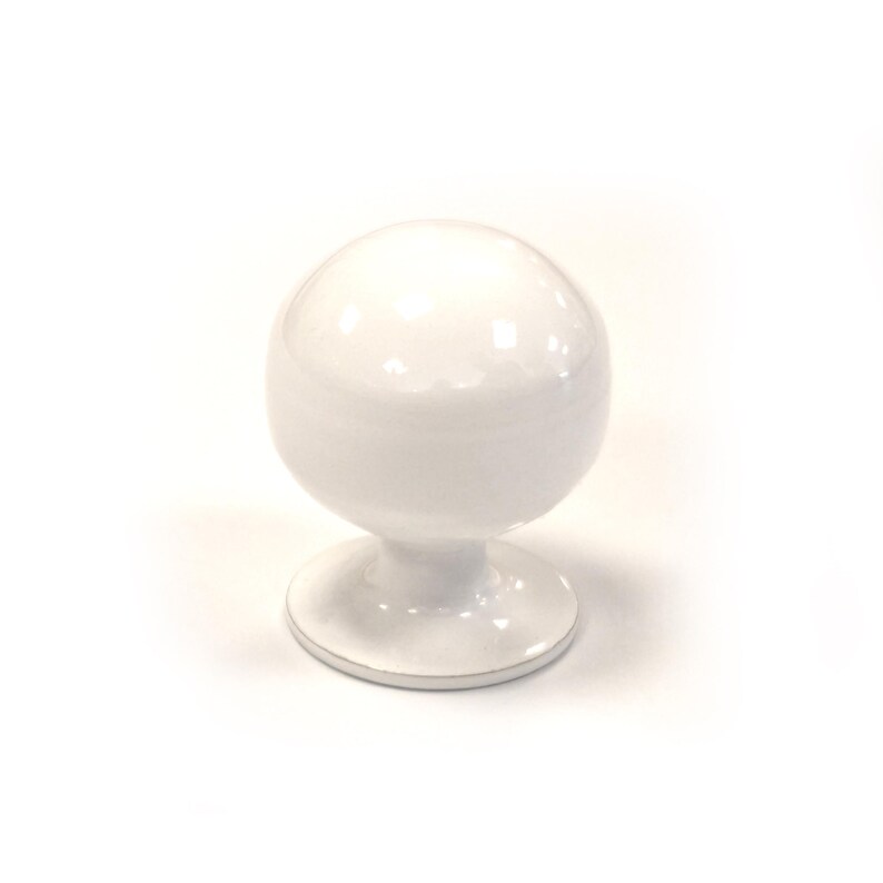 May include: A white, spherical cabinet knob with a glossy finish. The knob sits atop a short, rounded stem and a circular base. The design is simple and modern, suitable for various furniture styles. The knob is made of ceramic or porcelain.