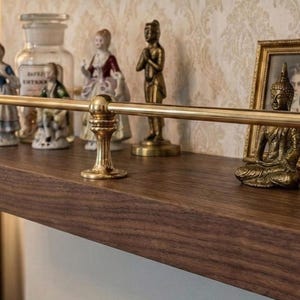 May include: A dark wood shelf with brass accents and decorative items. The shelf holds framed portraits, figurines, and a small Buddha statue. A brass bar runs along the front of the shelf, adding a touch of elegance.