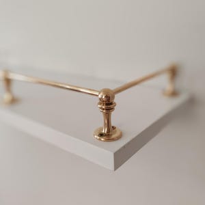 May include: A white shelf with a brass-colored metal railing. The railing has a rounded design and is attached to the shelf with decorative supports. The shelf is a simple, modern design.