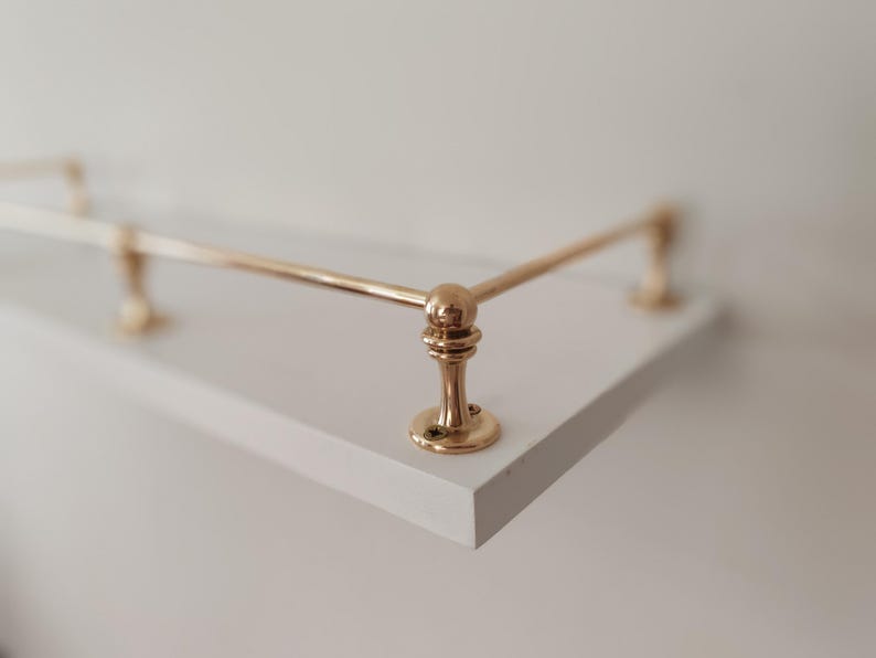 May include: A white shelf with a brass-colored metal railing. The railing has a rounded design and is attached to the shelf with decorative supports. The shelf is a simple, modern design.