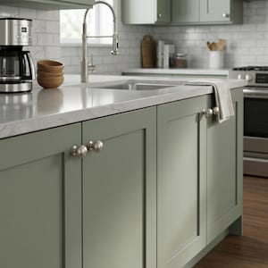 Peut inclure: Îlot de cuisine moderne avec des armoires vert sauge et des poignées en nickel brossé. Le comptoir est gris clair avec un motif marbré. Un robinet et une cafetière en acier inoxydable sont visibles, ainsi que des bols en bois.