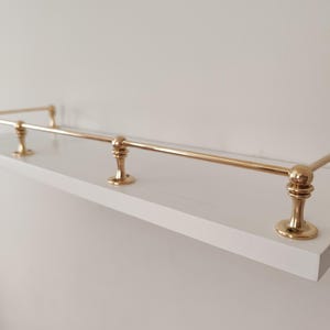 May include: A white shelf with brass-colored metal railing. The shelf has a rectangular shape and is mounted on a white wall. The brass railing has decorative, rounded supports.