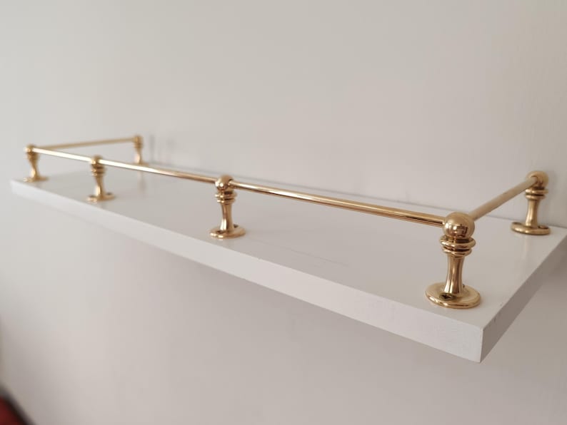 May include: A white shelf with brass-colored metal railing. The shelf has a rectangular shape and is mounted on a white wall. The brass railing has decorative, rounded supports.