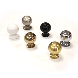 May include: Seven spherical cabinet knobs in different finishes are displayed. The knobs are in white, silver, black, gold, and chrome. Each knob has a round base and a smooth, polished surface, suitable for furniture hardware.