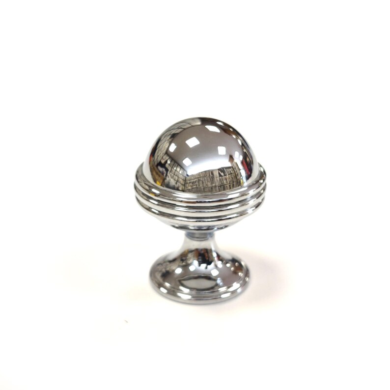 May include: A shiny, chrome-finished cabinet knob with a spherical top and a pedestal base. The spherical top has a reflective surface, and the base has a circular design. The knob is designed for furniture and home decor.