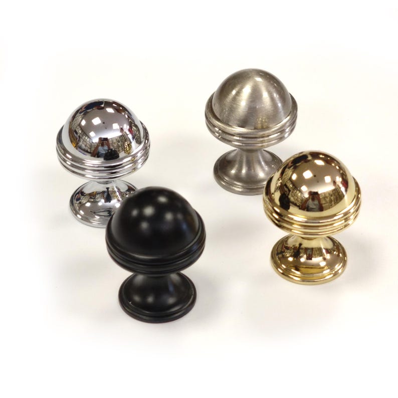May include: Four decorative cabinet knobs in different finishes: chrome, brushed nickel, black, and gold. Each knob features a spherical top with a ridged band and a pedestal base. The knobs are displayed on a white surface.