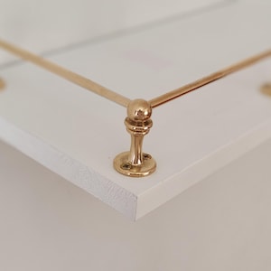 May include: A white shelf with a gold-colored metal railing. The railing has decorative ball-shaped accents and is attached to the shelf with round, flat bases. The shelf's edge is visible, showing a textured surface.