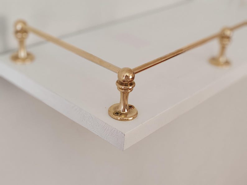 May include: A white shelf with a gold-colored metal railing. The railing has decorative ball-shaped accents and is attached to the shelf with round, flat bases. The shelf's edge is visible, showing a textured surface.