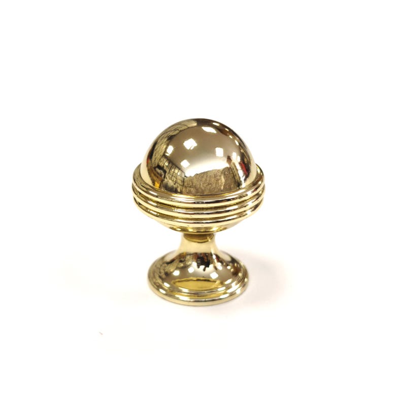 May include: A gold-colored cabinet knob with a spherical top and a decorative, ridged band. The knob sits on a pedestal base. The hardware is designed for furniture and home decor.