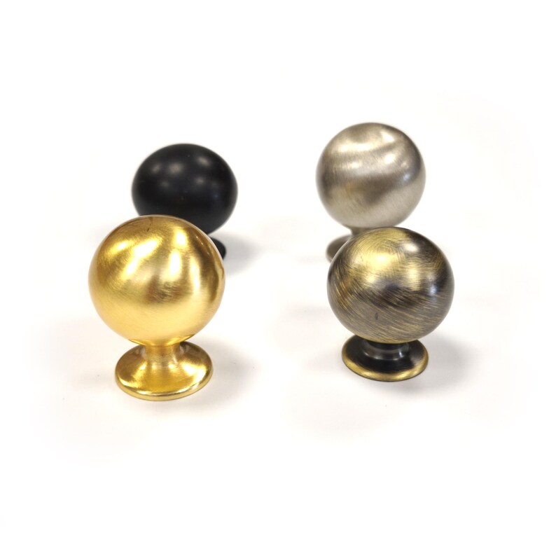 Op de afbeelding: Vier decoratieve kastknoppen in verschillende metallic afwerkingen. De knoppen zijn bolvormig met een kleine basis. Afwerkingen zijn onder meer goud, zwart, zilver en een bronsachtige kleur. De knoppen worden tegen een witte achtergrond weergegeven.