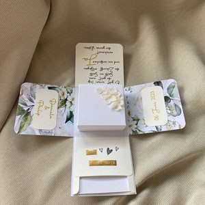 Könnte beinhalten: Eine weiße Box mit floralem Design und dem Text "Daniela & Philip" und "06. Juni 2023". Die Box ist geöffnet und enthält eine weiße Karte mit dem Text "You" und "Alles Gute" in goldener Schrift.