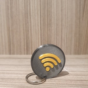 Op de afbeelding: Een grijze en gouden sleutelhanger met een wifi-symbool. De sleutelhanger is gemaakt van plastic en heeft een metalen ring.