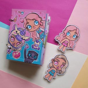 May include: A small pink binder with cartoon illustrations of a girl, a unicorn, and a cat. Two stickers of the girl are next to the binder. The background is pink, white, and yellow.