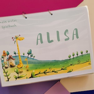 Könnte beinhalten: Ein farbenfroher illustrierter Buchumschlag mit einer Giraffe und einem kleinen Haus. Der Titel des Buches lautet "mein erstes Spielbuch" und der Name "ALISA" ist in großen grünen Buchstaben geschrieben.
