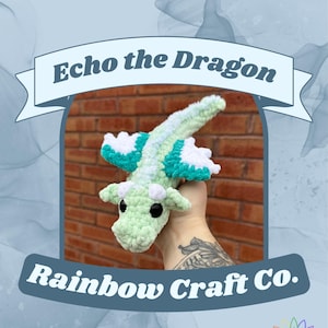 以下が含まれることがあります： 薄緑色、白、ターコイズ色の手編みドラゴンのおもちゃ。 ドラゴンは大きな黒い目と白い翼のディテールが特徴です。 ドラゴンの上には「Echo the Dragon」、下には「Rainbow Craft Co.」と書かれたテキストがあります。