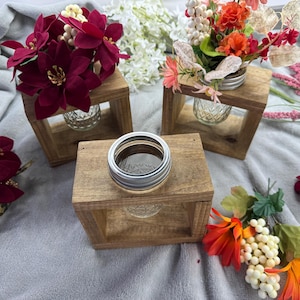 May include: Wooden candle holders with glass inserts, each holding a decorative floral arrangement. The flowers are in shades of red, orange, and white. A mason jar ring sits atop a similar wooden block.