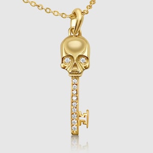 Peut inclure: Un collier avec un pendentif en forme de clé en or, orné d'un motif de crâne et d'accents de cristal transparent. Le crâne présente un design détaillé avec un nez et des orbites saillants.