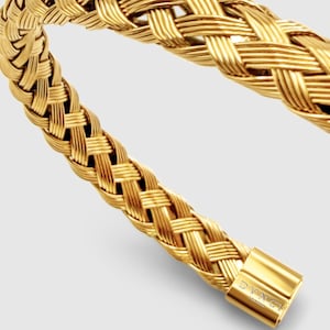 May include: A gold-colored bracelet featuring a woven, braided metal design. The bracelet has a smooth, polished cylindrical clasp at the end with the text DVNCI LONDON engraved on the surface. The metal has a bright, reflective finish throughout.