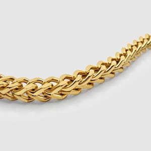 Può includere: Una collana a catena color oro caratterizzata da un fitto motivo a maglie intrecciate a spiga. Le maglie metalliche sono lucidate a specchio, creando una struttura continua, testurizzata e flessibile che si curva con una finitura brillante.