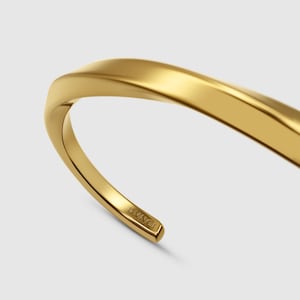 May include: A gold-toned metal cuff bracelet with a smooth, polished finish and a curved, open-ended design. The interior end of the bracelet features the engraved text DVNCI, showing a clean, reflective surface and a rounded, tapered profile.