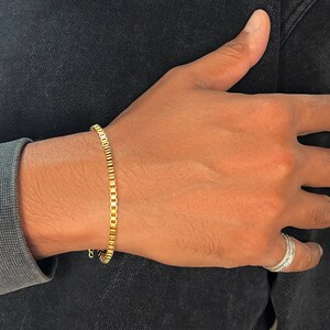 May include: A gold chain bracelet with a square link design. The bracelet is worn on a person's wrist.
