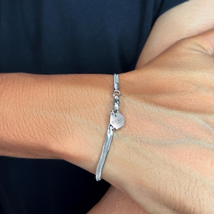May include: A silver bracelet with a snake chain design. The bracelet features a small, hexagonal charm with the word "DVNCI" engraved on it. The bracelet is secured with a lobster clasp.