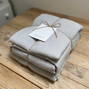 May include: A stack of light grey linen bedding is wrapped in twine and tied with a bow. A white tag with the text "melkhomeware" is attached to the twine.