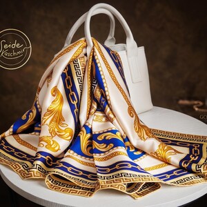 May include: A white handbag with a draped silk scarf. The scarf has a cream background with gold and blue ornate patterns. The design includes chain links, Greek key borders, and floral accents. The scarf is draped over the handbag on a white table.
