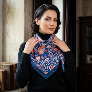 May include: A square silk scarf featuring a navy blue border and a colourful floral design in orange, pink, and white against a blue backdrop. The scarf is worn around the neck, highlighting its detailed pattern and colour scheme.