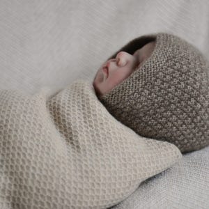 Könnte beinhalten: Ein Baby trägt eine braune Strickmütze mit Wabenmuster, eingewickelt in eine beige Strickdecke mit Wabenmuster.