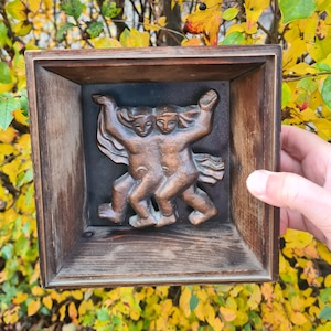 Op de afbeelding: Een houten lijst met een gesneden houten beeldhouwwerk van twee dansende figuren erin. Het beeldhouwwerk is in bronskleur geverfd.