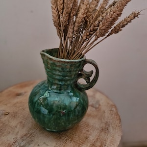 Op de afbeelding: Een kleine, groene keramische kruik met een handvat, gevuld met gedroogde tarwearen. De kruik heeft een glanzende afwerking en staat op een houten oppervlak. De tarwe heeft een lichtbruine kleur, die contrasteert met de groene tint van de kruik.