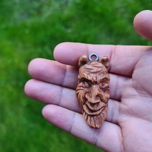May include: A wooden pendant carved in the shape of a face with a mischievous expression. The pendant has a loop at the top for hanging.