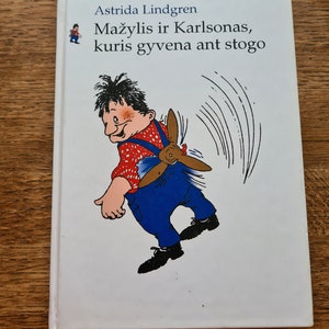 Può includere: Una copertina di libro bianca con il titolo "Astrid Lindgren, Mažylis ir Karlsonas, kuris gyvena ant stogo" in testo nero. La copertina presenta un'illustrazione di un ragazzo con un'elica sulla schiena.