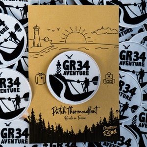 Peut inclure: Un patch rond blanc avec une broderie noire représentant un randonneur sur un sentier avec le texte "GR34 AVENTURE". Le patch est entouré d'autres patchs avec le même design, et une carte brune avec un dessin d'un phare, du soleil et des nuages. La carte porte également le texte "Patch thermocollant Brodé en France" et "Capitaine Rémi".