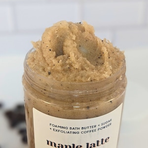 Op de afbeelding: Close-up van een bruine suikerscrub met koffiedik in een glazen pot. Het etiket vermeldt "Foaming Bath Butter + Sugar + Exfoliating Coffee Powder" en "maple latte".