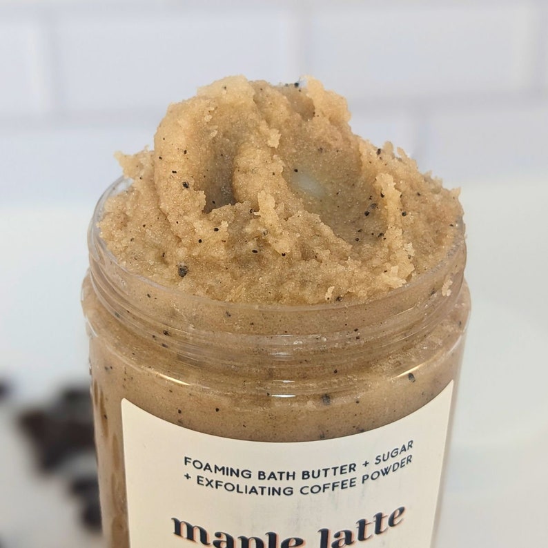 Op de afbeelding: Close-up van een bruine suikerscrub met koffiedik in een glazen pot. Het etiket vermeldt "Foaming Bath Butter + Sugar + Exfoliating Coffee Powder" en "maple latte".