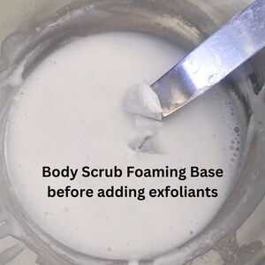 Op de afbeelding: Een close-up van een witte, schuimende body scrub-basis in een metalen kom. Een spatel tilt een deel van de basis uit de kom. De tekst "Body Scrub Foaming Base before adding exfoliants" staat onder de kom.