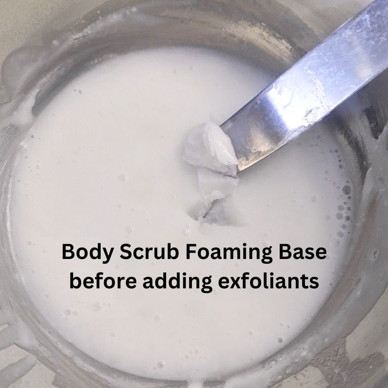 Op de afbeelding: Een close-up van een witte, schuimende body scrub-basis in een metalen kom. Een spatel tilt een deel van de basis uit de kom. De tekst "Body Scrub Foaming Base before adding exfoliants" staat onder de kom.
