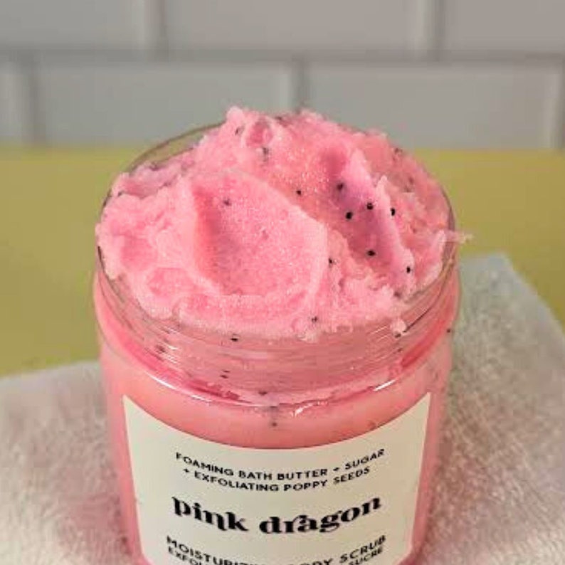 Op de afbeelding: Roze badboter met papaverzaad in een glazen pot met een roze etiket waarop staat "FOAMING BATH BUTTER + SUGAR + EXFOLIATING POPPY SEEDS pink dragon MOISTUR EXFOL LOY SCRUB CRE".
