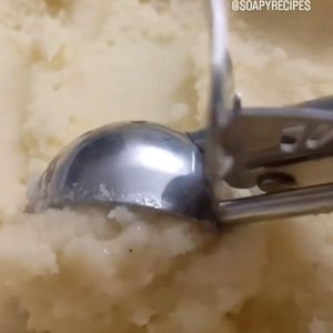 Op de afbeelding: Een close-up van een zilveren ijsbollepel die een grote kom met aardappelpuree opschept. De lepel is gedeeltelijk ondergedompeld in de puree, en de steel is zichtbaar op de achtergrond. De tekst "@SOAPYRECIPES" is zichtbaar in de rechterbovenhoek van de afbeelding.