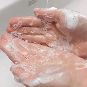 Op de afbeelding: Close-up van twee handen bedekt met witte zeepschuim die worden afgespoeld onder een straal water in een witte gootsteen.