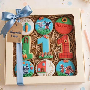 Op de afbeelding: Een vierkante geschenkdoos met een doorzichtig venster bevat negen versierde koekjes. De koekjes tonen stripfiguren, het cijfer één in verschillende stijlen en de naam Alessandro. Een blauw satijnen lint en een bruin label met de tekst Alessandro 1 anno zijn aan de doos bevestigd.