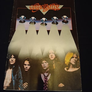 以下が含まれることがあります： エアロスミスのアルバムカバー。カバーには、スポットライト付きの5つのダイヤモンドのグラフィックの下にバンドメンバーがいます。バンド名は、黒い背景にダイヤモンドの上にオレンジ色のスクリプトで書かれています。