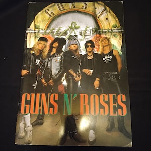 以下が含まれることがあります： ガンズ・アンド・ローゼズのアルバムカバー。革ジャンとサングラス姿のメンバーが写っています。バンド名は大きなオレンジと緑の文字で表示されています。背景には、スカルと時計のデザインが施されています。