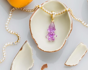 May include: A gold-toned necklace with a clear, purple gnome pendant. The necklace is set with small, clear stones. The gnome pendant is in a white, shell-shaped dish. The image also includes a yellow object and pieces of a broken shell.