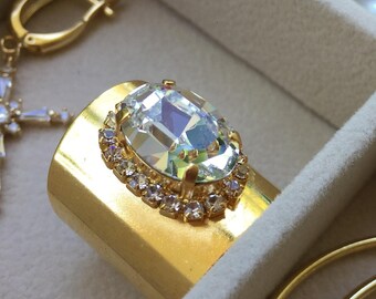 Könnte beinhalten: Ein goldfarbener Ring mit einem großen, klaren, oval geschliffenen Edelstein in der Mitte, umgeben von kleineren, klaren Steinen. Der Ring ist in einem goldenen Band gefasst. Im Hintergrund sind weitere goldene Schmuckstücke zu sehen.